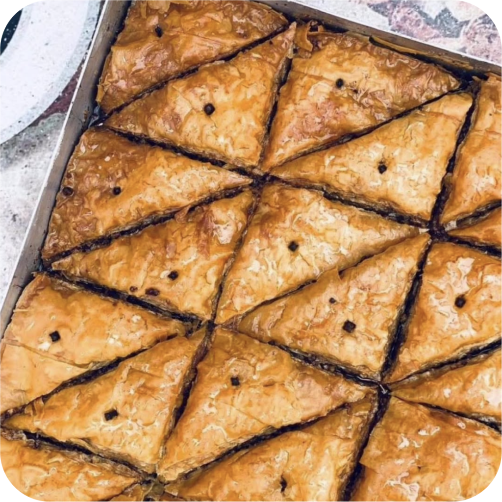 Freshly baked baklava cut into triangular pieces in a tray. The golden-brown pastry glistens with syrup, with visible layers and a rich, sweet aroma.