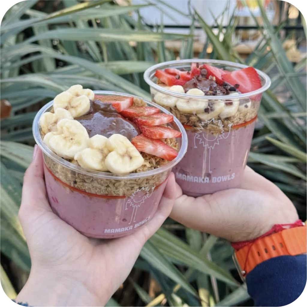 Two hands hold acai bowls topped with banana slices, strawberries, granola, and chocolate. The background features green foliage.