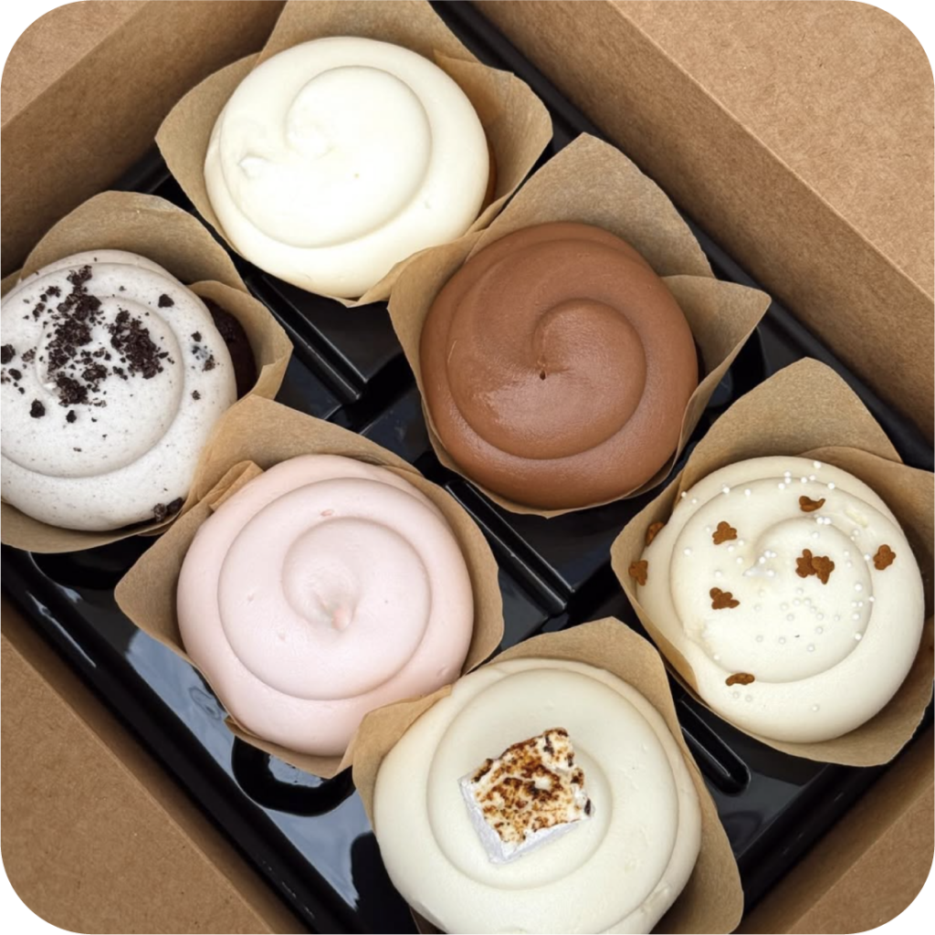 A box of six gourmet cupcakes, each with unique toppings, including chocolate, cream, sprinkles, and a toasted marshmallow, displayed on a black tray.