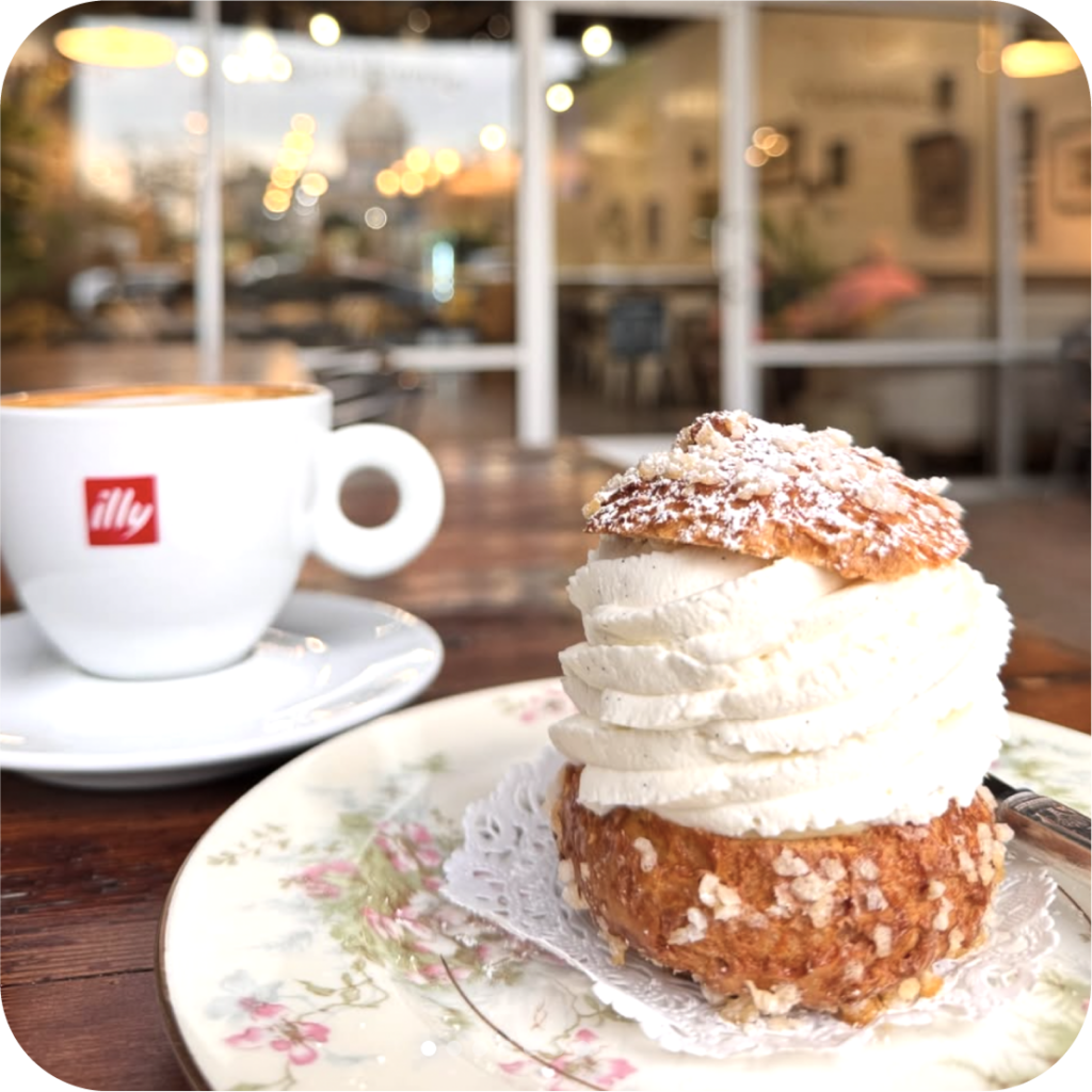 A cappuccino in an Illy cup sits on a wooden table next to a cream-filled pastry on a floral plate. A cozy café ambience is blurred in the background.