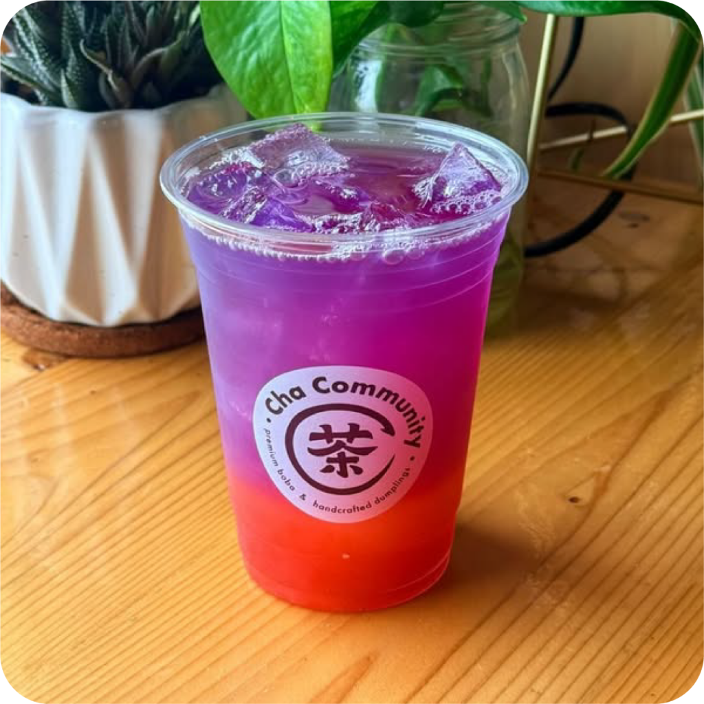 A vibrant purple and red gradient iced drink in a clear cup with "Cha Community" logo, sits on a wooden table next to leafy plants. Refreshing and inviting.