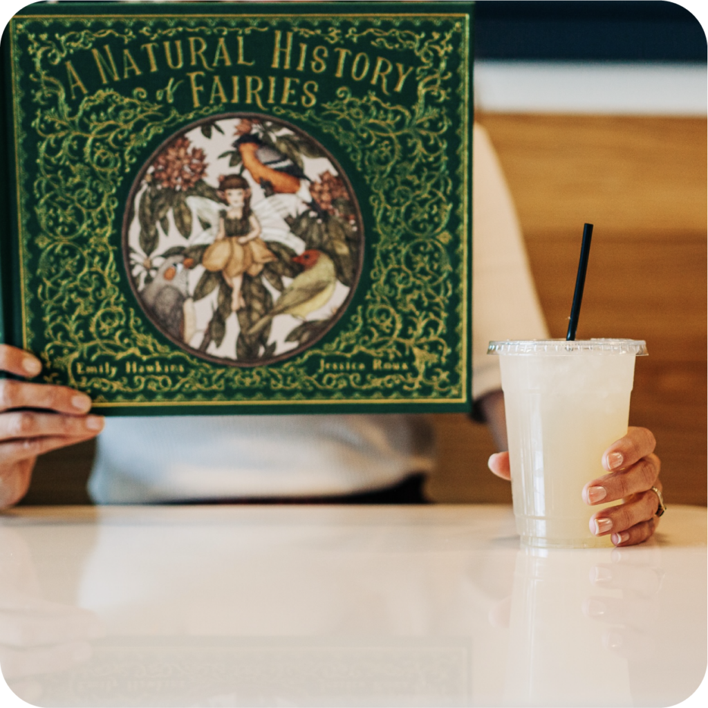 A person reads "A Natural History of Fairies," with an ornately illustrated green cover, while holding a cold drink in a clear cup with a black straw.