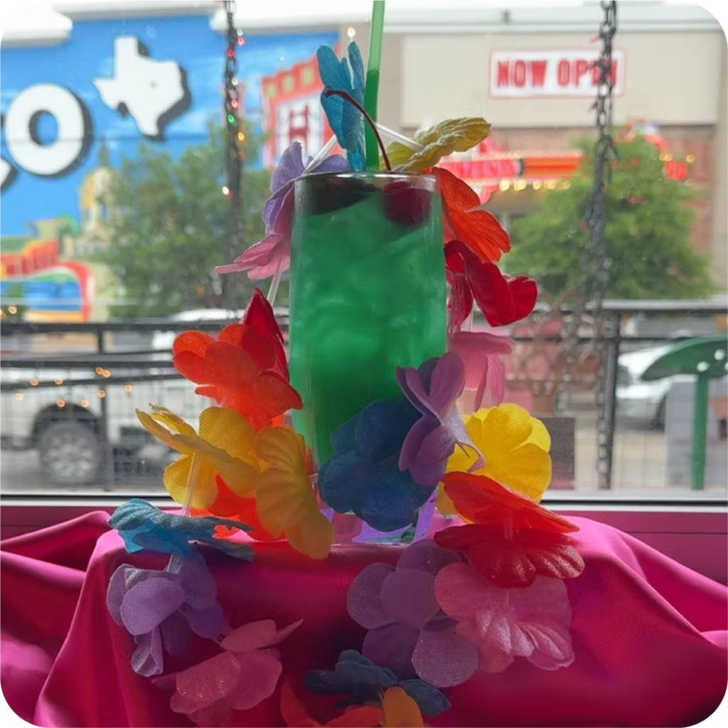 A vibrant green cocktail topped with a cherry and straw, surrounded by a colorful lei, sits on a magenta cloth. A mural and building are visible in the background.