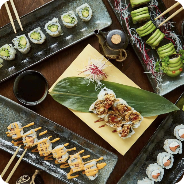 Overhead view of a colorful sushi spread with rolls, avocado, sauces, chopsticks, tea, and decorative greenery on dark plates.