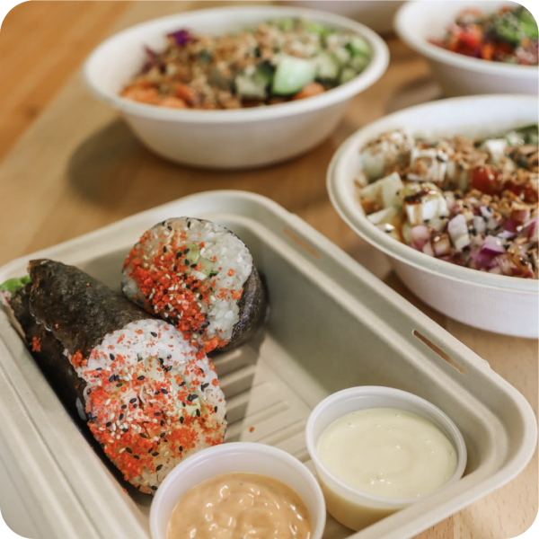 A tray of sushi rice rolls with colorful sesame toppings and dipping sauces, surrounded by fresh salad bowls on a wooden table.