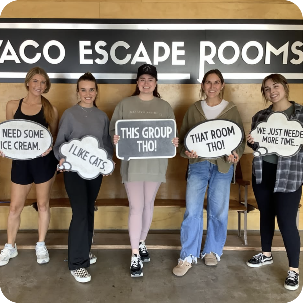 Five smiling women pose at Waco Escape Rooms, holding humorous speech-bubble signs about ice cream, cats, the room, and needing more time.