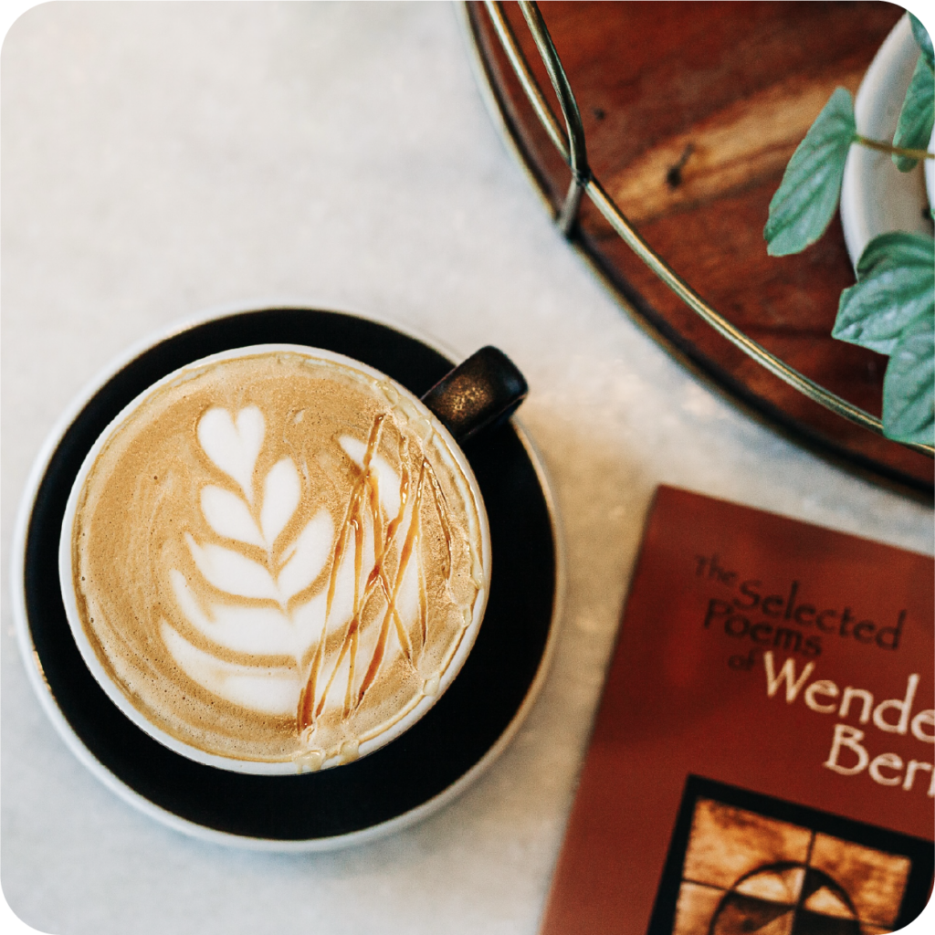 Top-down view of a latte with leaf-shaped foam art beside a red Wendell Berry poetry book and leafy plant, creating a cozy café mood.