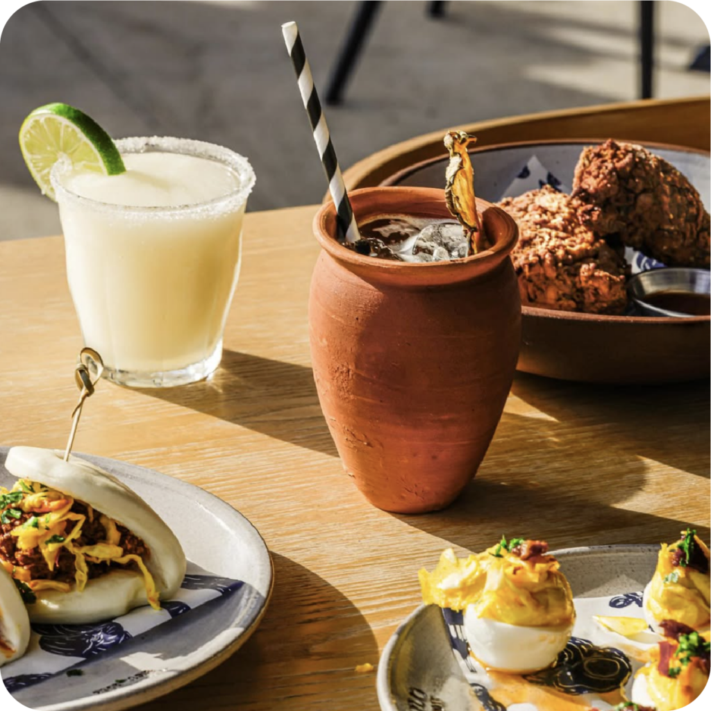 Sunlit outdoor table with a lime-garnished creamy drink, clay cup cocktail, tacos, and fried bites; warm, inviting dining scene.