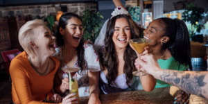 Four friends laugh and toast at a lively bar; one wears a tiara while holding a cocktail, capturing a joyful birthday celebration.